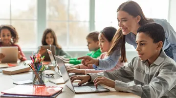 Kinder lernen am Tisch mit Laptops und Tablets. Eine Lehrerin hilft und zeigt auf den Bildschirm. Es gibt Stifte, Hefte und Fenster mit Licht.