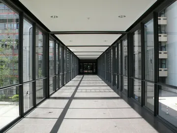 Langer Gang mit vielen Fenstern. Die Sonne scheint hinein und macht Schatten auf den Boden. Draußen sieht man Bäume und Gebäude.