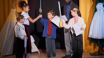 Kinder spielen Theater auf der Bühne mit gelben Vorhängen. Ein Kind mit Krone hält ein Schwert. Ein Erwachsener schaut zu. Zelte und Kostüme sind im Hintergrund.