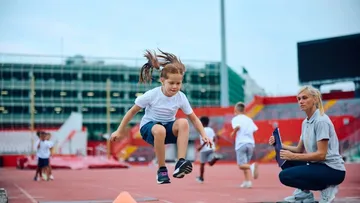 Ein Mädchen macht Weitsprung, während die Trainerin zusieht.