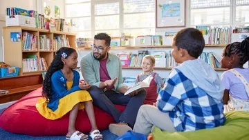 Ein Mann liest Kindern in einer Bücherei vor. Alle sitzen gemeinsam auf Sitzkissen und hören gespannt zu.