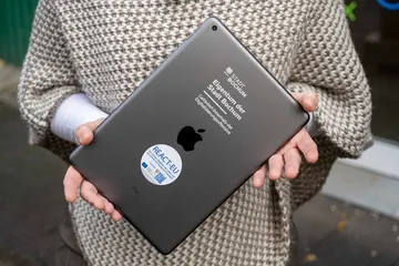 Eine Person hält ein schwarzes iPad mit der Rückseite nach vorne in beiden Händen. Auf dem Gerät sind mehrere Aufdrucke zu sehen: das Apple-Logo, der Hinweis „Eigentum der Stadt Bochum – Gefördert innerhalb der Digitalisierungsmaßnahme“ sowie ein runder Aufkleber mit dem EU-Programm „REACT-EU“. Die Person trägt einen grob gestrickten Poncho in Beige-Braun-Tönen über einem weißen Langarmshirt.