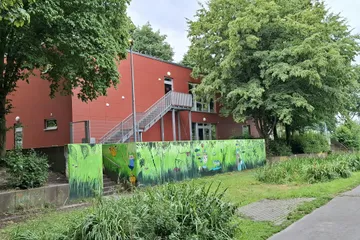 Blick auf den Neubau im hinteren Schulbereich, genutzt für Sprachförderung und Differenzierung, im Vordergrund des Bildes natürlich bepflanzte Rigolen zur Entsiegelung des Schulhofs und Versickerungshilfe