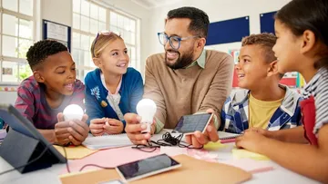 Sieben Personen arbeiten in einem hellen Büro mit Pflanzen und Fenstern. Sie haben Laptops, Notizbücher und Haftnotizen auf den Tischen. Sie tragen bunte Kleidung.