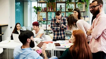 Drei Leute sitzen an einem Holztisch und reden. Eine trägt ein gestreiftes Hemd, eine ein rotes T-Shirt und eine ein blaues Hemd. Auf dem Tisch liegt ein Notizbuch. Im Hintergrund gibt es Bücherregale und bunte Wände.