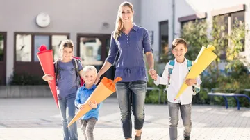 Eine Mutter geht mit drei Kindern vor einer Schule. Die Kinder tragen Rucksäcke und bunte Schultüten (rot, orange, gelb). Alle lächeln und sind lässig gekleidet. Die Schule hat große Fenster und eine Uhr. Die Sonne scheint.