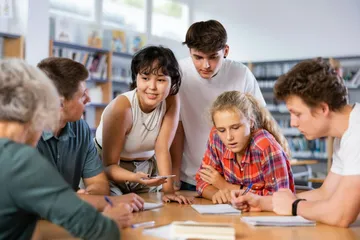 Jugendliche lernen zusammen am Tisch in der Bibliothek. Sie halten Stifte und Notizbücher. Eine Frau zeigt auf ein Blatt. Es gibt Bücherregale und Fenster mit Licht.