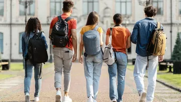 Fünf junge Leute gehen über einen Weg vor einem großen Gebäude. Sie tragen Rucksäcke und lässige Kleidung wie Jeans und T-Shirts. Das Gebäude hat viele Fenster und sieht alt aus. Die Sonne macht die Szene hell und freundlich.