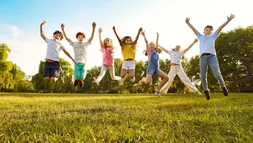 Ein Kind mit langen Haaren holt lächelnd ein Buch aus einem Regal. Es trägt ein beiges Kleid und steht auf einem grünen Teppich. Im Hintergrund gibt es Kissen und Bücher.