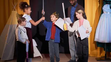Kinder spielen Theater auf der Bühne mit gelben Vorhängen. Ein Kind mit Krone hält ein Schwert. Ein Erwachsener schaut zu. Zelte und Kostüme sind im Hintergrund.