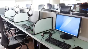 Ein Büroarbeitsplatz mit mehreren Schreibtischen. Jeder Tisch hat einen Stuhl, einen Computerbildschirm, eine Tastatur, eine Maus und Kopfhörer. Die Schreibtische sind durch kleine Wände getrennt. Große Fenster lassen viel Licht herein. Der Boden ist mit dunklen Fliesen bedeckt.