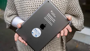 Eine Person hält ein schwarzes iPad mit der Rückseite nach vorne in beiden Händen. Auf dem Gerät sind mehrere Aufdrucke zu sehen: das Apple-Logo, der Hinweis „Eigentum der Stadt Bochum – Gefördert innerhalb der Digitalisierungsmaßnahme“ sowie ein runder Aufkleber mit dem EU-Programm „REACT-EU“. Die Person trägt einen grob gestrickten Poncho in Beige-Braun-Tönen über einem weißen Langarmshirt.
