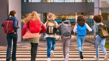 Sechs lachende Kinder rennen über einen Zebrastreifen vor einer Schule. Sie tragen bunte Rucksäcke und lässige Kleidung wie Jeans und Jacken. Ihre Haare wehen im Wind. Die Schule hat große Fenster. Die Sonne macht die Szene hell und fröhlich.