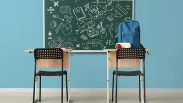 Ein leeres Klassenzimmer mit einem Tisch und zwei Stühlen vor einer beschriebenen Tafel. Ein Rucksack, ein Apfel und Bücher liegen auf dem Tisch. Die Wände sind blau, ein Fenster lässt Licht herein.